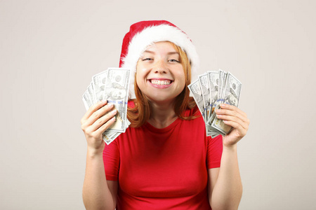 金钱作为圣诞礼物的概念。 幸福的年轻红头发的女人的肖像，持有一束100美元的，作为新年礼物。 灰色孤立的背景复制空间关闭。