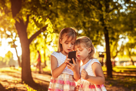 两个年轻漂亮的金发姐妹在日落时在户外使用手机玩得很开心的肖像。