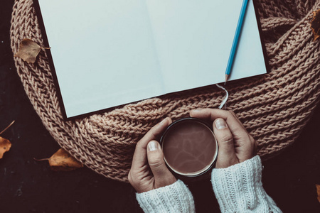 在棕色针织围巾和秋叶的背景上，用铅笔在女性手中和记事本上放一杯热可可。