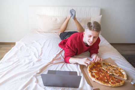 一个快乐的年轻人躺在一个明亮舒适的房间里的床上，用笔记本电脑上网，吃披萨。 周末看电影和披萨