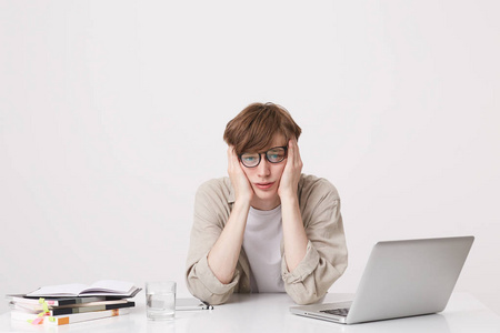 一个悲伤而不快乐的年轻学生戴着眼镜，穿着米色衬衫，坐在桌子旁看着沮丧，笔记本电脑和笔记本电脑被隔离在白色背景上
