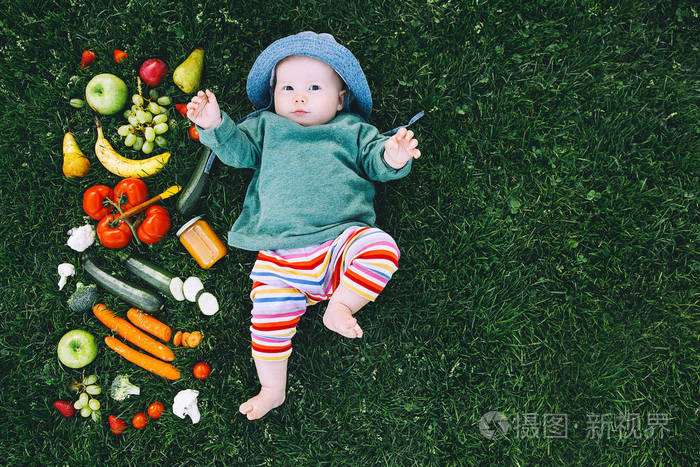 婴儿穿着五颜六色的衣服，尝试不同新鲜水果和蔬菜的食物和框架，在绿草背景的顶部。 健康儿童营养婴儿喂养。