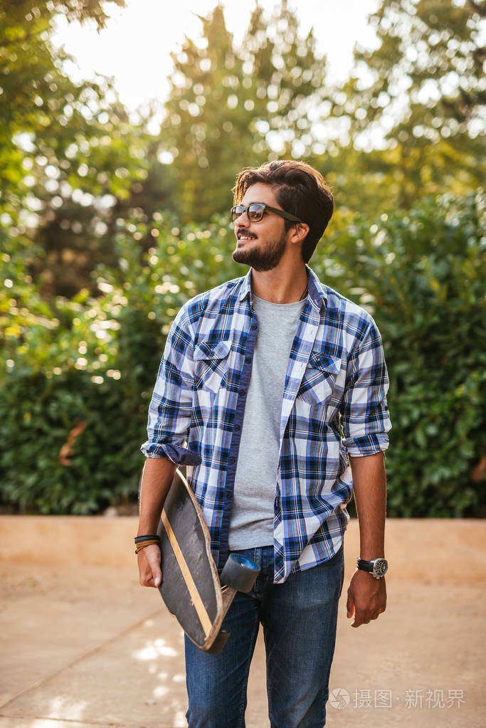 英俊迷人的男人拿着长板带着太阳镜走在街上