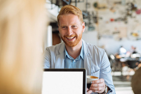 一位年轻微笑的商人坐在咖啡馆里一边喝咖啡一边工作的肖像。
