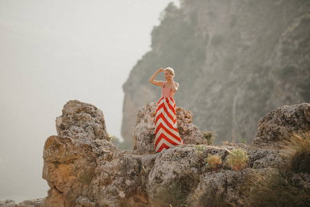 一位穿着红色连衣裙的迷人金发女士在悬崖顶上跳舞，背景是山。 山景。 西班牙的晚上