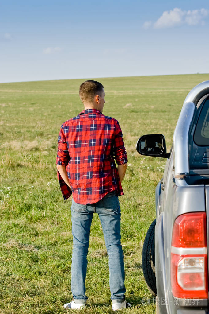 穿着红色格子衬衫的年轻人背靠在绿野上的汽车旁