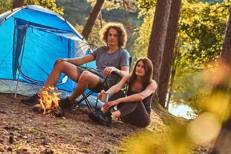 年轻的夫妇在阳光明媚的日子里, 在森林里露营的篝火旁取暖。