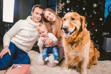 主题圣诞节和新年家庭圈和家庭宠物。 爸爸妈妈和一岁的白种人妇女坐在圣诞树附近的地板上，带着礼物和大狗饲养拉布拉多金毛猎犬。