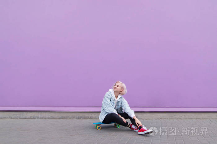 时尚快乐的女孩坐在滑板上靠在紫色的墙上微笑。一个漂亮的金发女孩穿着红色运动鞋坐在滑板上的紫色背景的肖像。