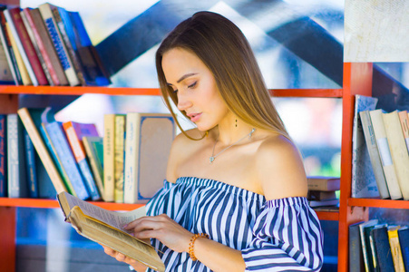 在现代图书馆明亮多彩的书架旁阅读教科书的年轻女子