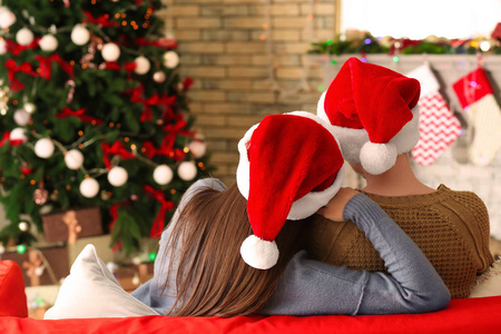 可爱的年轻夫妇戴着圣诞帽坐在家里的沙发上
