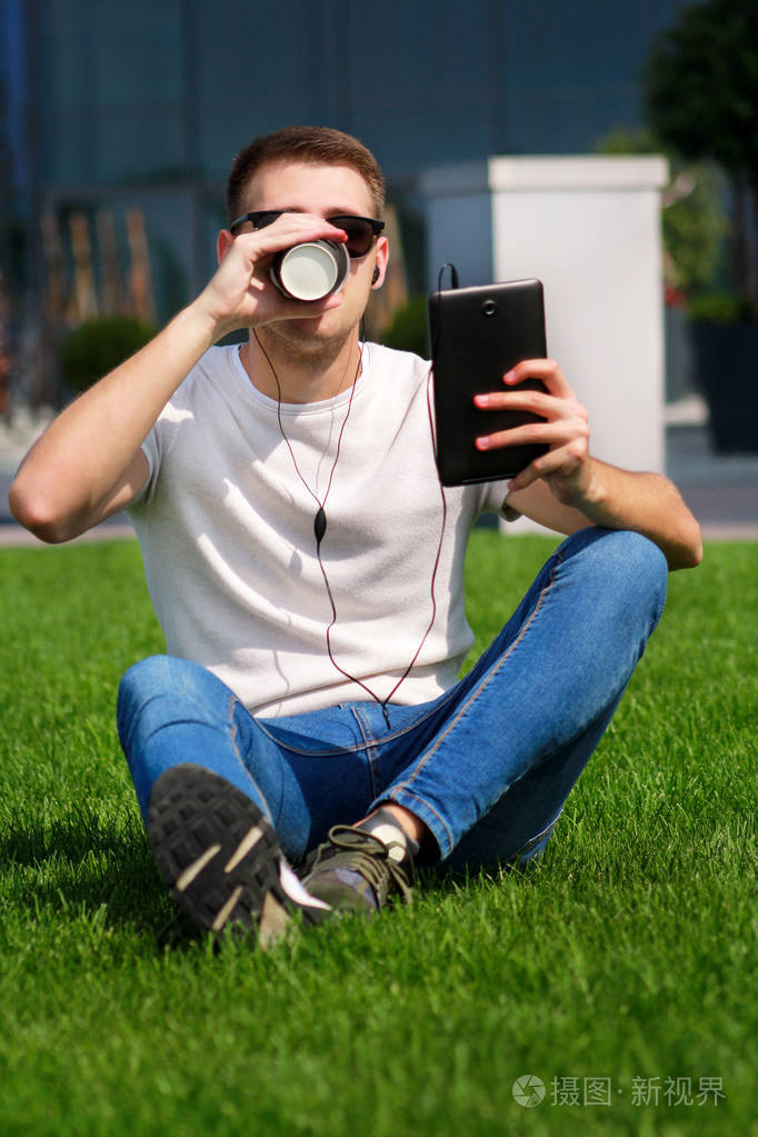 年轻的帅哥带着太阳镜,用平板电脑和耳机喝咖啡,坐在草地上,享受现代