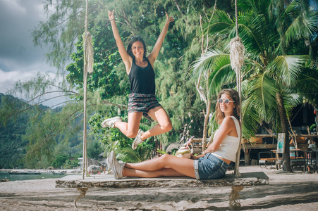 两个漂亮的女朋友在海滩上享受秋千和喝椰子。 热带旅游假期