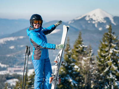 一位微笑的女子滑雪者站在滑雪场的山顶上，指着她的科普空间周围令人惊叹的自然冬季景观，享受快乐娱乐季节性运动