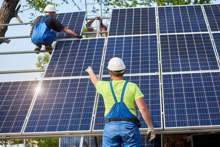 男性工程师技术人员站在未完成的高外部太阳能电池板照片Voltaic系统前面的后视图，两名工人在高平台上指向闪亮的大蓝色表面。