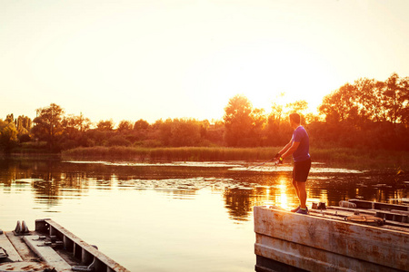 日落时站在桥上钓鱼的年轻人.快乐的费瑟曼享受夏天的爱好照片