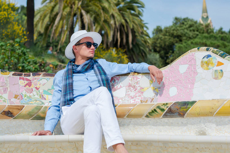 一个戴着帽子太阳镜围巾的年轻人坐在公园的长凳上，盖尔建筑师加迪巴塞罗那西班牙。