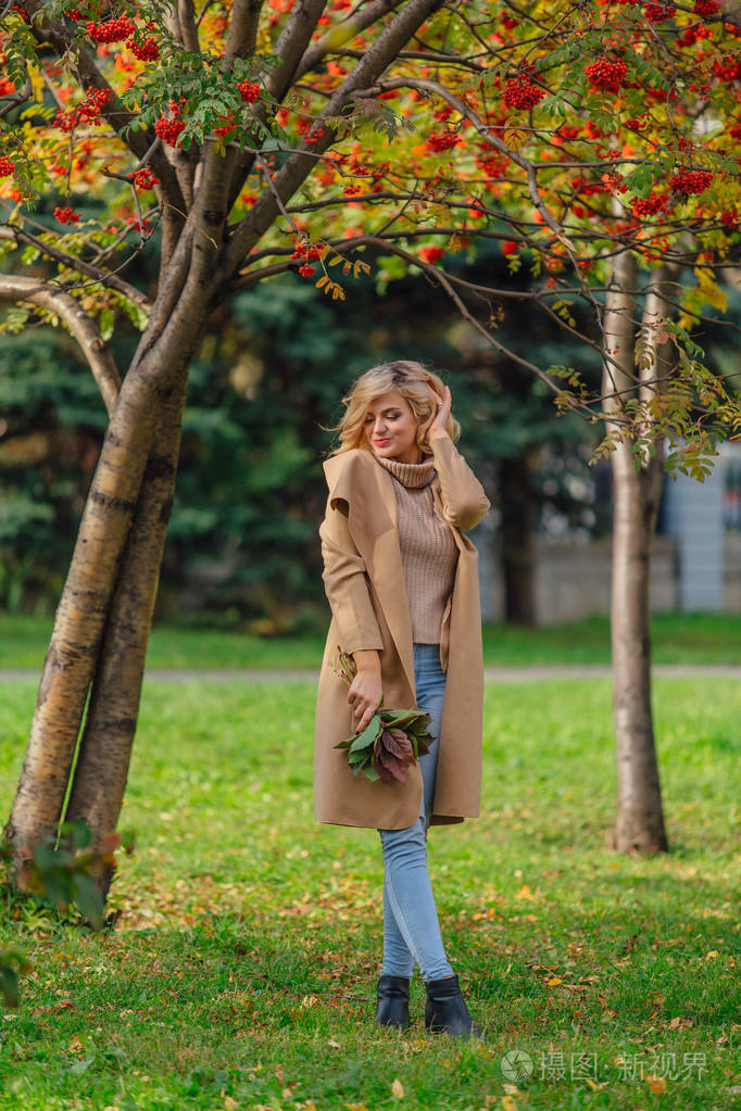 美丽优雅的金发女人穿着外套,站在罗旺树下,手里拿着一束罗旺莓和秋天