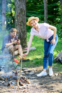 女孩在稻草帽子烹调食物在篝火自然背景。露营技巧。每个露营者都应该知道篝火烹饪的秘诀。简单的提示, 以改善您的篝火烹饪。在棍子上烹