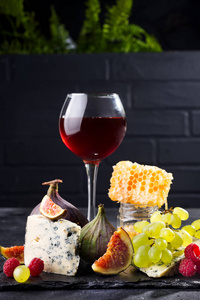 葡萄, 乳酪, 无花果和蜂蜜用红色的杯子在石头黑色背景之上在砖黑色墙壁与绿色花, 拷贝空间