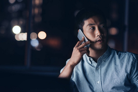 年轻的亚洲商人深夜在办公室工作时用手机说话，背景是城市灯光