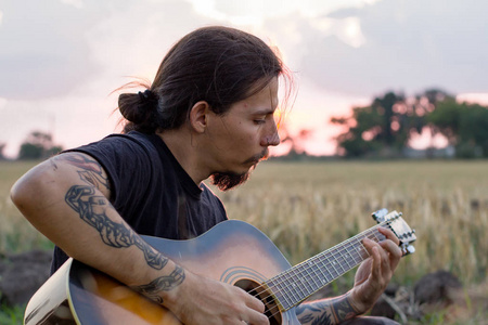 在美丽的夕阳下，年轻的纹身男性在夏天的田野里玩声吉他