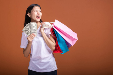 女人拿着购物袋和现金孤立在橙色背景下。 年轻的亚洲女人穿着白色T恤，心情愉快。 假日购物概念。