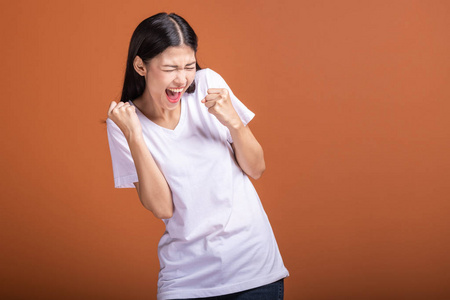 获胜的女人被隔离在橙色背景上。 年轻的亚洲时髦女人穿着白色T恤，大赢尖叫姿势两只手举起。 成功的年轻潮人概念。