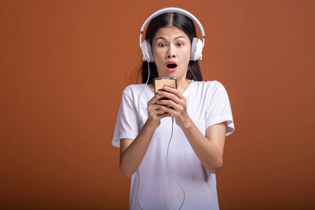 女人听音乐用电话隔离在橙色背景上。 年轻的亚洲女人带着手机听音乐，浏览音乐，心情很震惊。 时尚生活方式概念