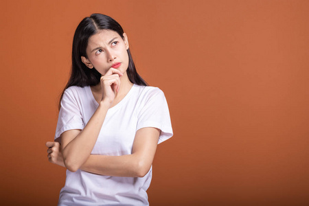 思考女人的橙色背景。 年轻的亚洲时髦女人穿着白色T恤，专注思考，把手指放在下巴上。 年轻女人时髦的概念。