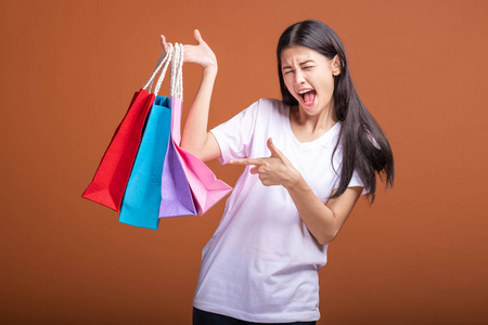 女人拿着购物袋，孤立在橙色背景下。 年轻的亚洲女人穿着白色T恤，高兴地指着袋子的心情。 购物概念。