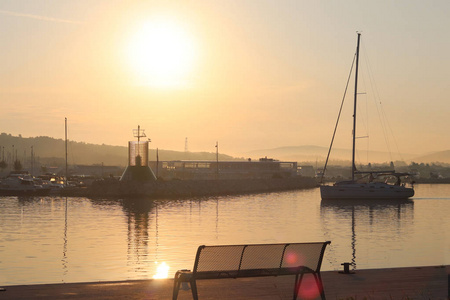 帆船在清晨的阳光下进入码头的马达下。 港口的黎明。 码头上的空凳靠在灯塔上。 返回和等待的符号。 平静的浪漫气氛。