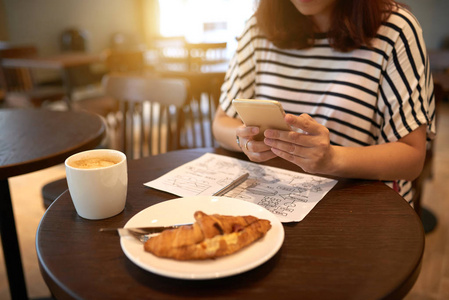 女性在咖啡桌上使用手机的部分景观，有一杯卡布奇诺和开胃的牛角面包