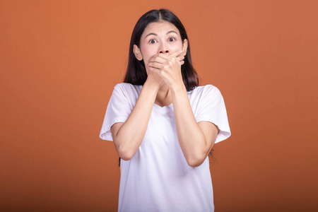 橙色背景下的惊喜女人。 年轻的亚洲时髦女人穿着白色T恤，摆着惊喜的姿势，两只手捂住嘴。 年轻女人时髦的概念。