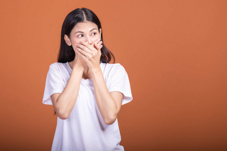 橙色背景下的惊喜女人。 年轻的亚洲时髦女人穿着白色T恤，摆着惊喜的姿势，两只手捂住嘴。 年轻女人时髦的概念。