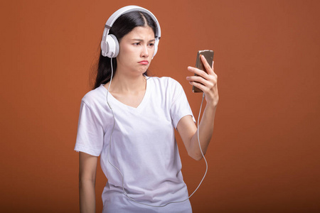 女人听音乐用电话隔离在橙色背景上。 年轻的亚洲女人带着手机听音乐，浏览音乐，心烦意乱。 时尚生活方式概念