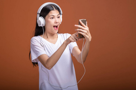 女人听音乐用电话隔离在橙色背景上。 年轻的亚洲女人带着手机听音乐，浏览音乐，兴奋的心情。 时尚生活方式概念