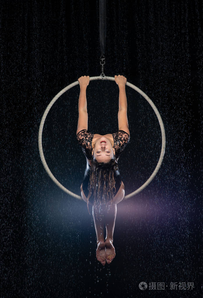 美丽的女杂技演员坐在空中箍的对称姿势在雨中黑色背景下