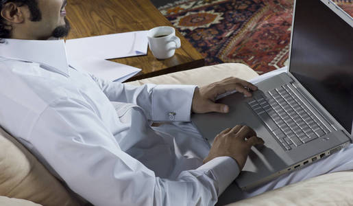 mideastern男人在家里用笔记本电脑，旁边桌子上放着一杯咖啡和文件