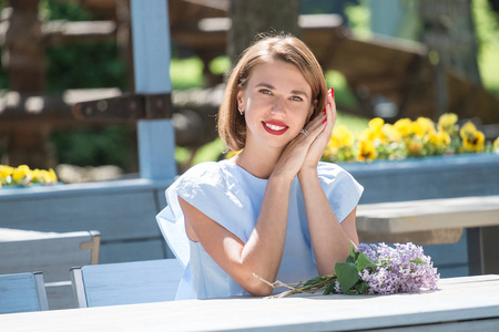一位年轻迷人的女士，身穿浅蓝色连衣裙，坐在夏日咖啡馆的桌旁，手捧一束清新的丁香花
