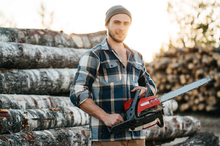 在锯木厂的背景下,身穿格子衬衫的大胡子伐木工人手里拿着电锯照片