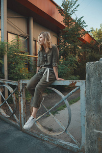 时髦的女孩在街上摆姿势，坐在一个弯曲的腿的钢制栅栏上。城市风格。街头时尚。