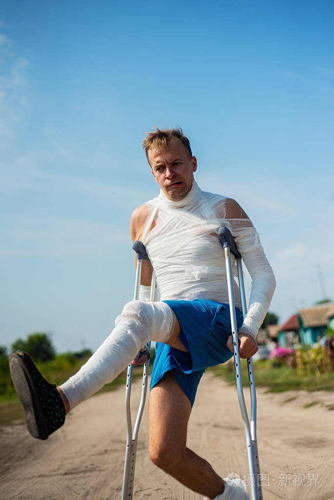 一个绑着绷带的年轻人拄着拐杖在乡间的路上散步玩得很开心做着鬼脸