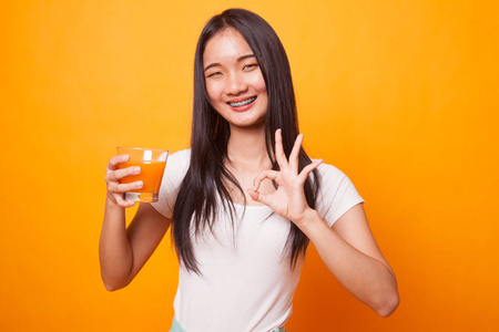 年轻的亚洲女人表演OK喝橙汁亮黄色背景