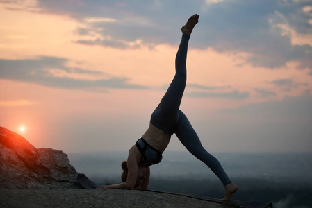 苗条的年轻体操女子在黄昏时在高岩石山上做复杂的有氧瑜伽练习的剪影，背景是美丽的深蓝色，多云的傍晚，夏日的天空和红色的夕阳。