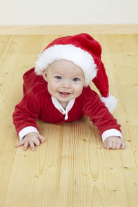 可爱的小宝宝穿着红色圣诞老人服装，微笑着看着相机，躺在木地板上