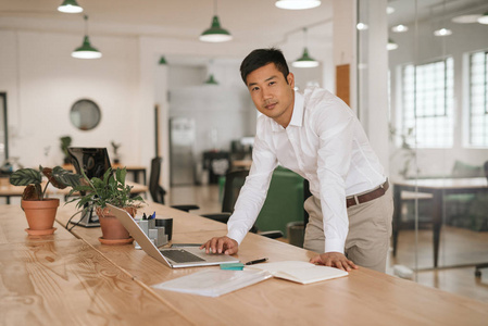 一位专注的年轻亚洲商人的肖像，他靠在一间现代办公室的桌子上，手提电脑在网上工作，还在看文件