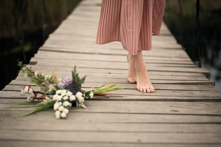 特写一个美丽的温柔女孩的腿，穿着一件桃色的连衣裙，走在农村的木桥上，地板上放着一束鲜花。