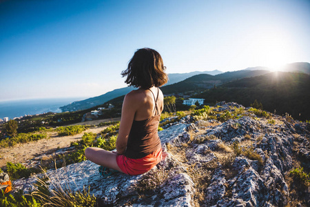 日落时，一个女孩坐在山顶上。 一个女人从事瑜伽，坐在莲花的位置。 冥想离文明很远。 漂亮的女背。 山和海的景色。