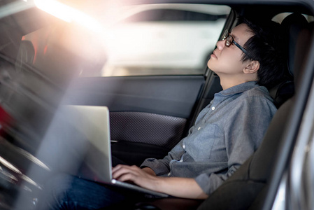 过度劳累的亚洲商人戴着眼镜，在车上驾驶座上用笔记本电脑工作时感到压力和疲劳。 男性企业家为他的事业努力工作。